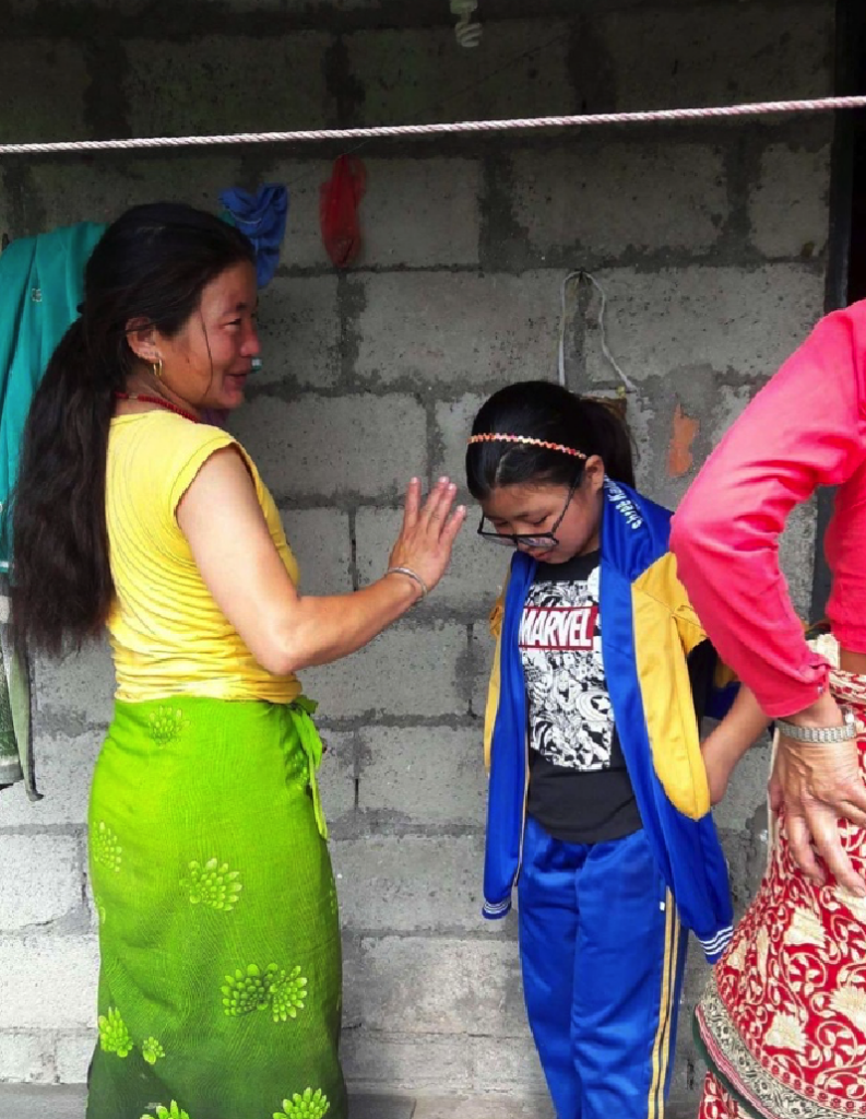 A Nepali woman and her daughter dressed in bright clothes, talk and laugh with each other in front of their breezeblock home
