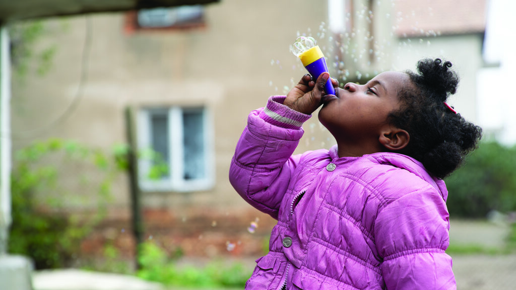 A little mixed race girl in a pink jacket blows bubbles into the air