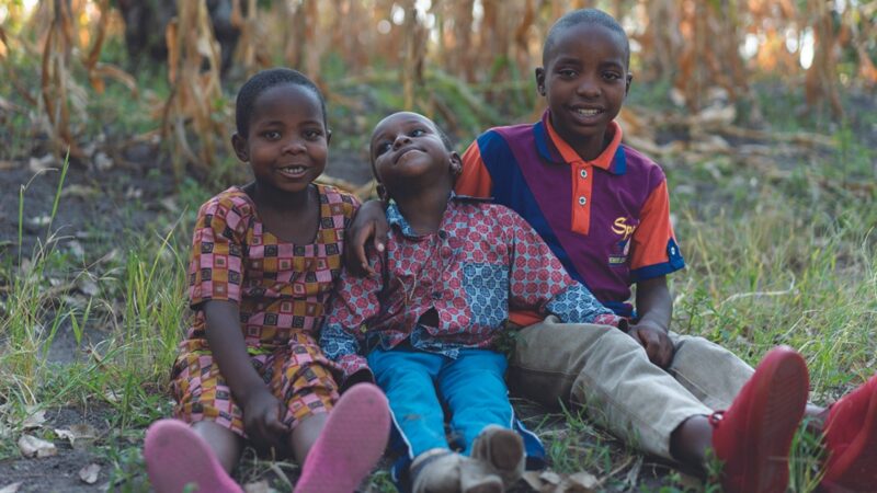 A young Rwandan boy, Carrol, sits on the ground between his win sister, Estrella, and his older brother, Eric.