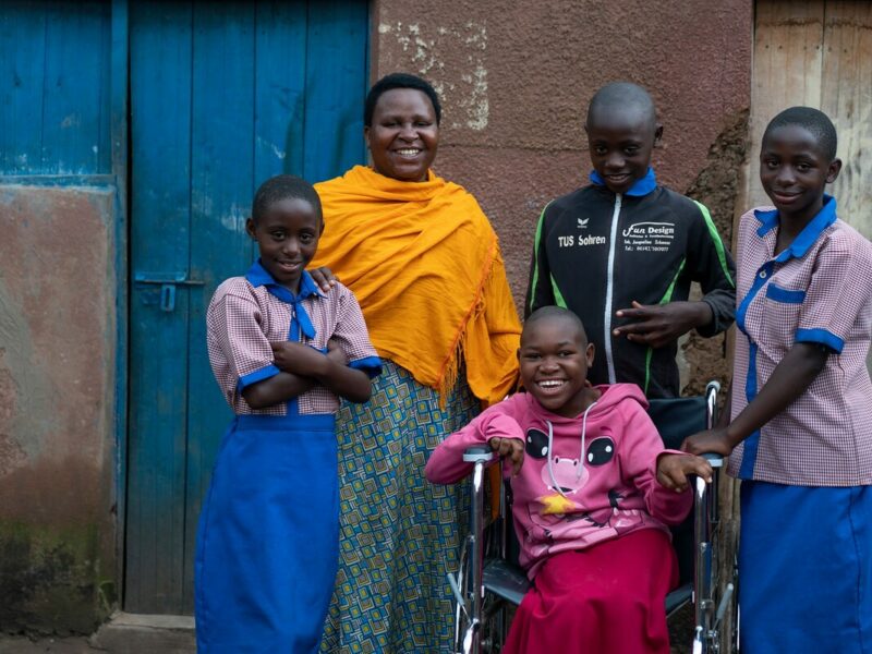 A young Rwandan girl, a wheelchair user, sits outside her house with her siblings and mother around her smiling