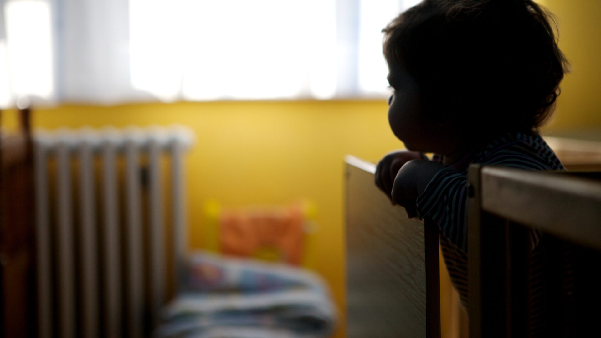 Silhouette of a small child leaning over the side of a cot in a dim room