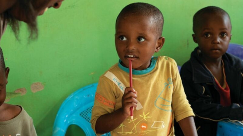 A young Rwandan girl in a yellow sweater stands behind a desk holding a pen, at a community hub.