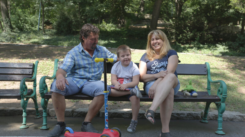 Georgi, who has down syndrome, sits on a bench in a park with his parents on either side and a red scooter. They are all laughing.