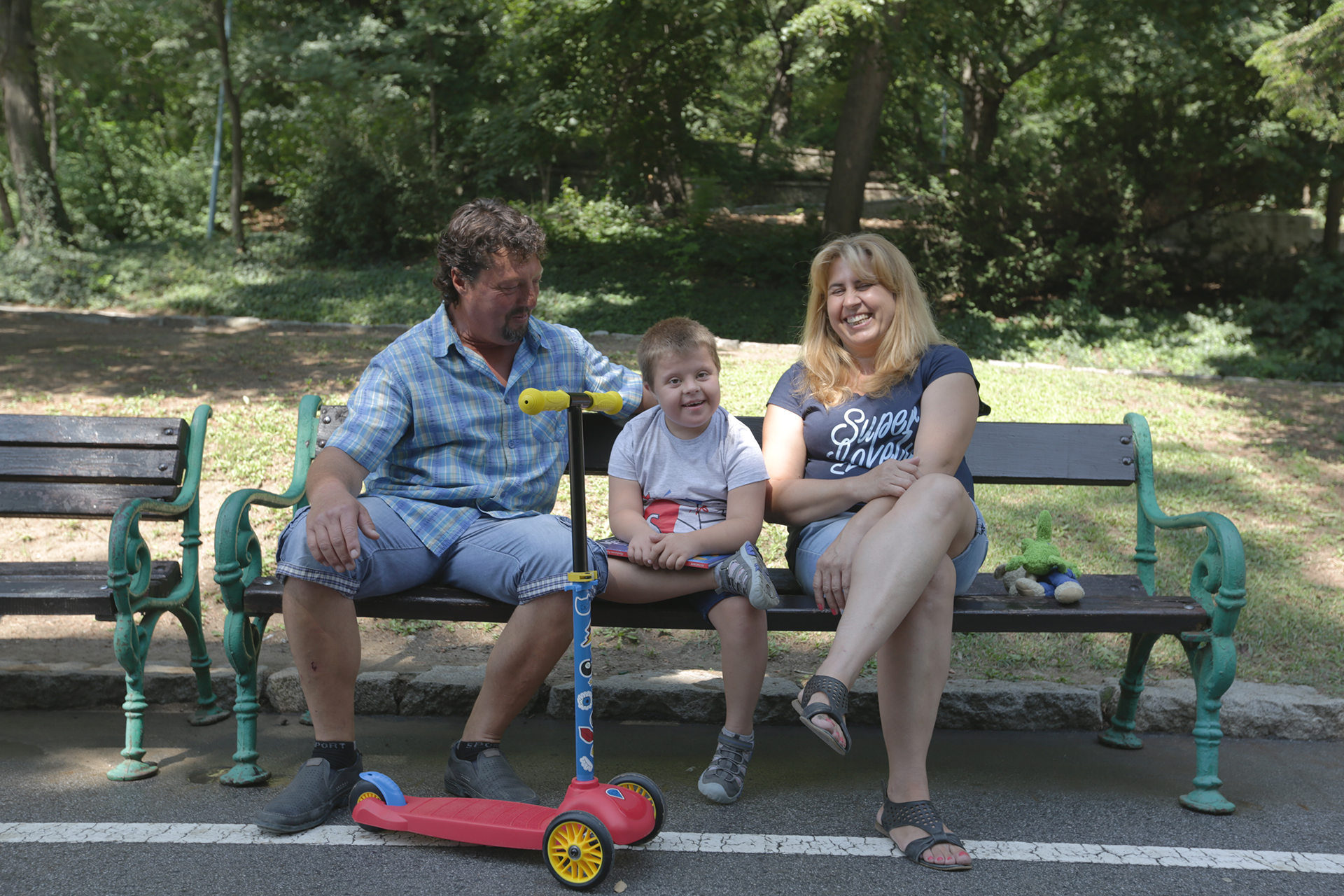 Georgi, who has down syndrome, sits on a bench in a park with his parents on either side and a red scooter. They are all laughing.