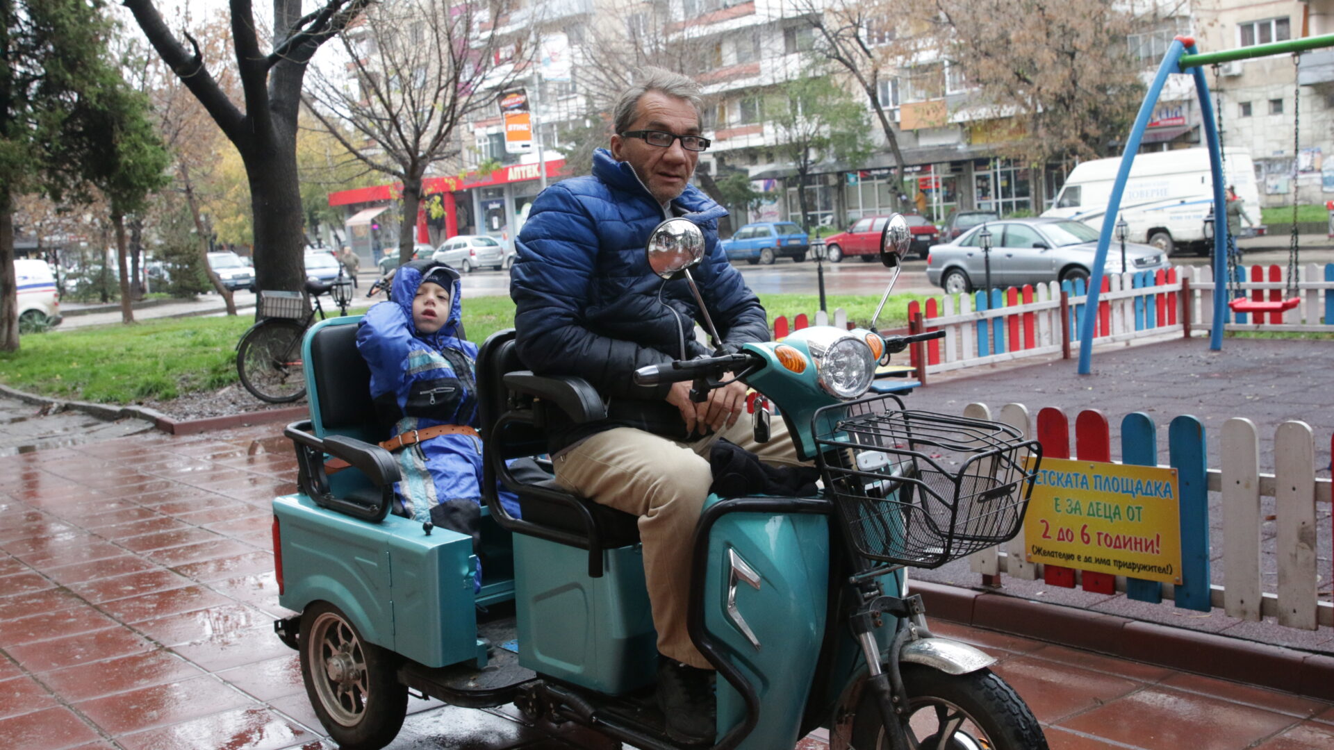 A father and his disabled son ride on a mobility scooter along a tree lined street in the rain