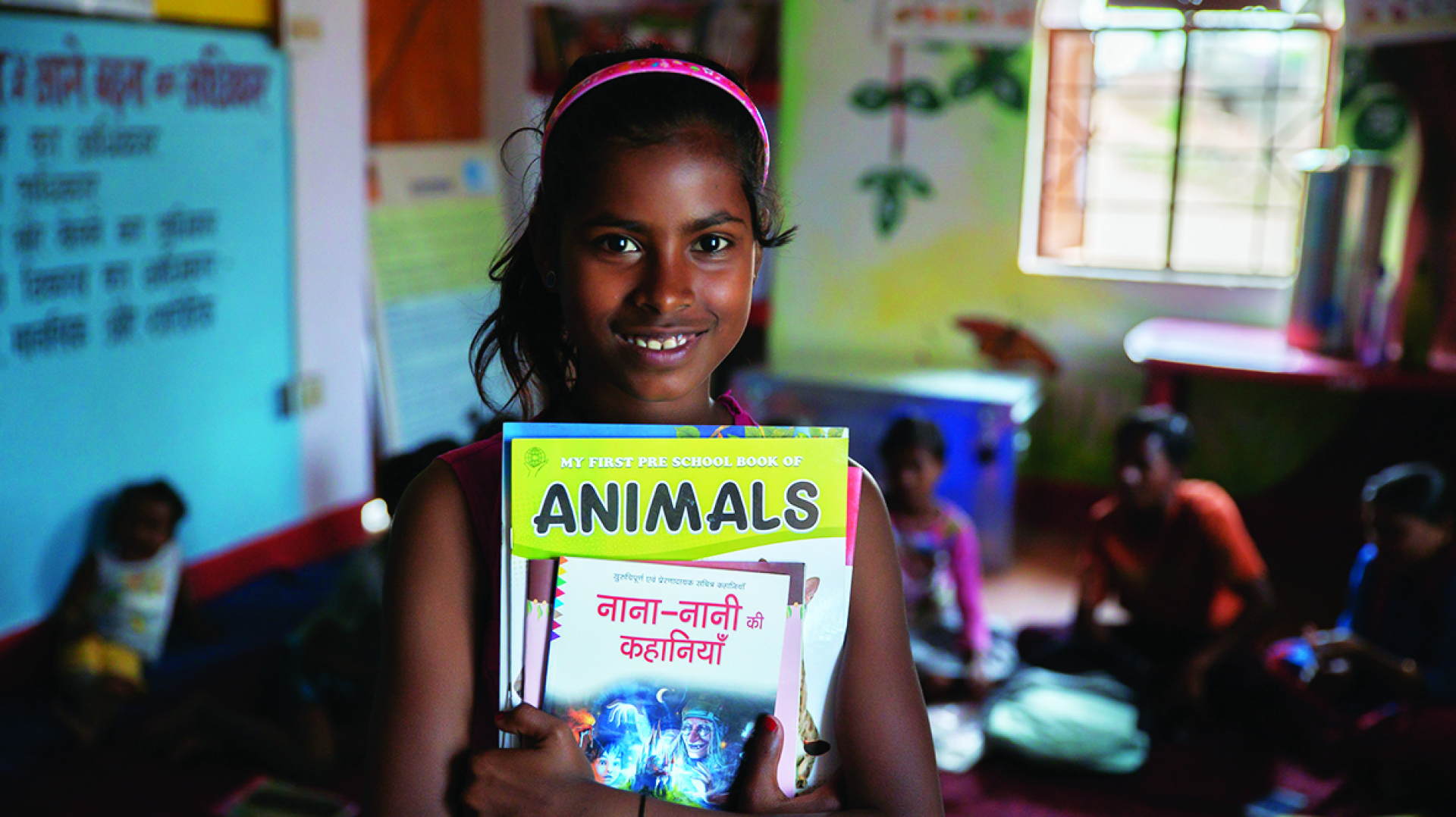 A young Indian girl stands in front of her classmates in the schoolroom, clutching her books and smiling at the camera