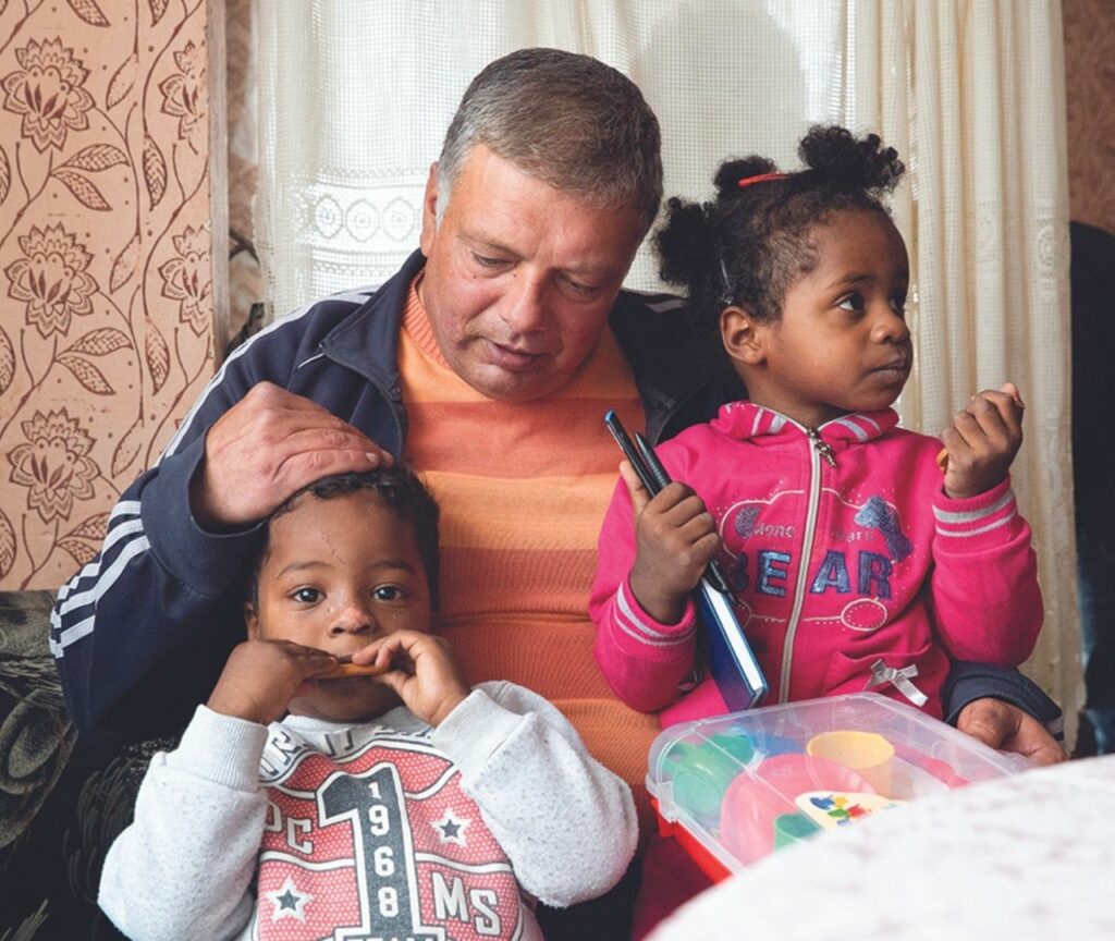 A middle aged white man with two mixed race children on his lap, sitting at a kitchen table