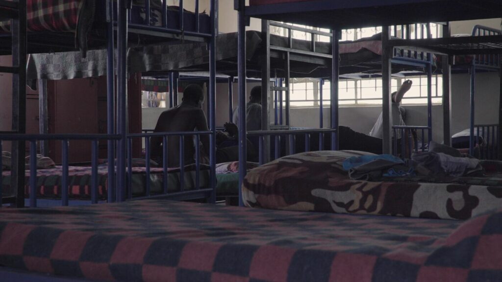 A child sits alone on the bottom bunk of a dim room filled with metal bunk beds, in an orphanage in Kenya