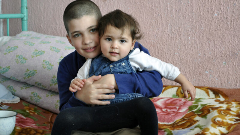 A young Moldovan boy holds his baby sister while they sit on the bed and smile at the camera