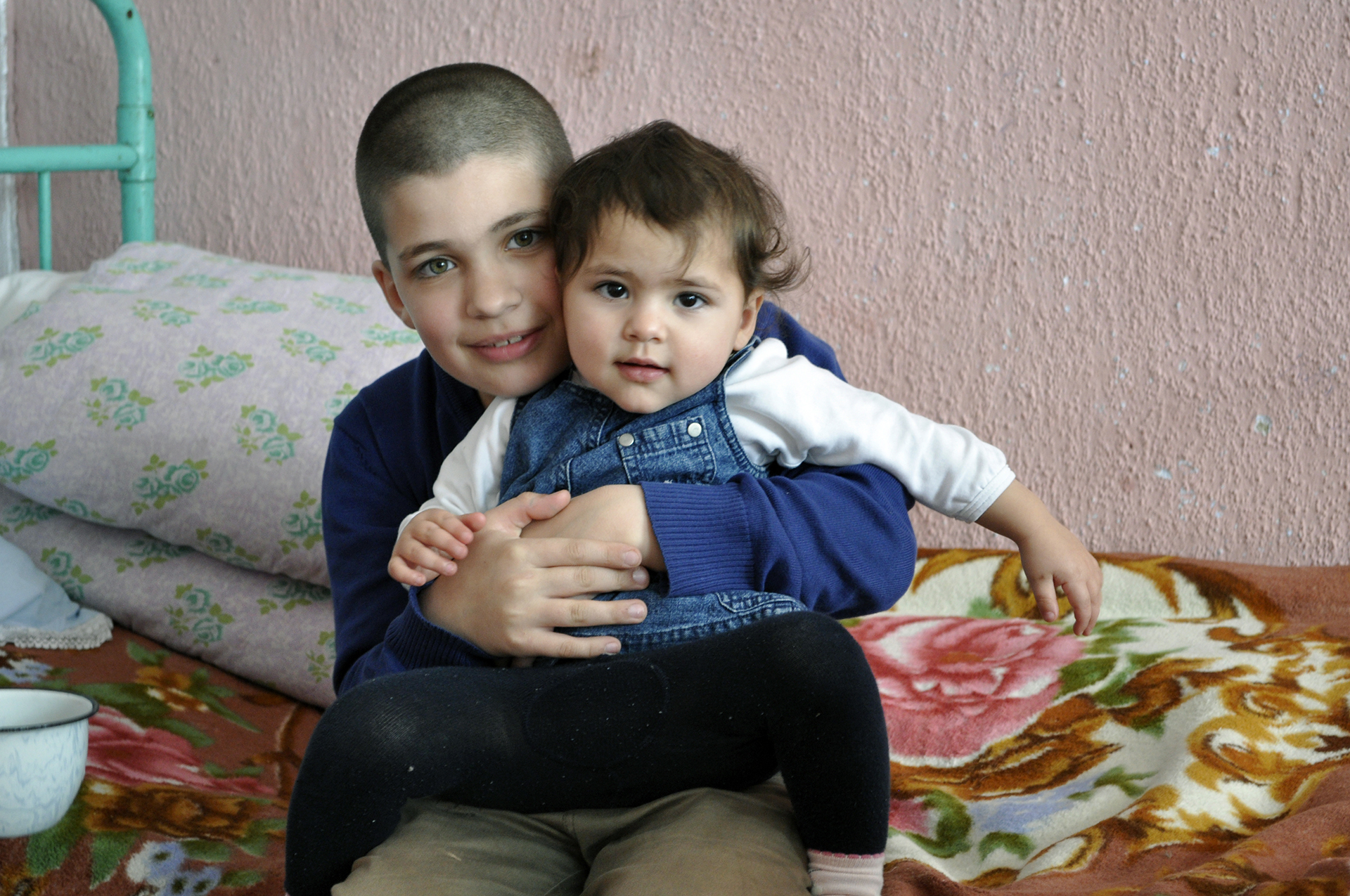 A young Moldovan boy holds his baby sister while they sit on the bed and smile at the camera