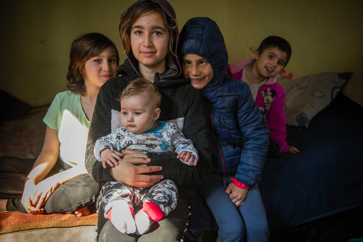 4 children and an older woman sit on the bed in a dark room, looking confidently at the camera