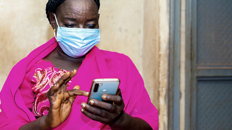 A Rwandan woman in a bright pink shawl and a PPE mask show from waist up - she's looking at her phone about to type