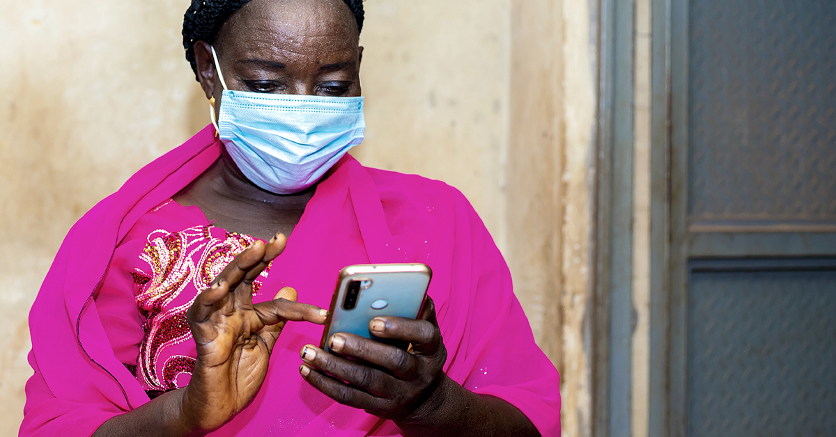 A Rwandan woman in a bright pink shawl and a PPE mask show from waist up - she's looking at her phone about to type