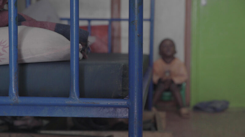 A child sits on the floor with his knees drawn up to his chest, by a blue metal bunk bed, in an orphanage in Kenya. The picture is blurred to preserve his anonymity.