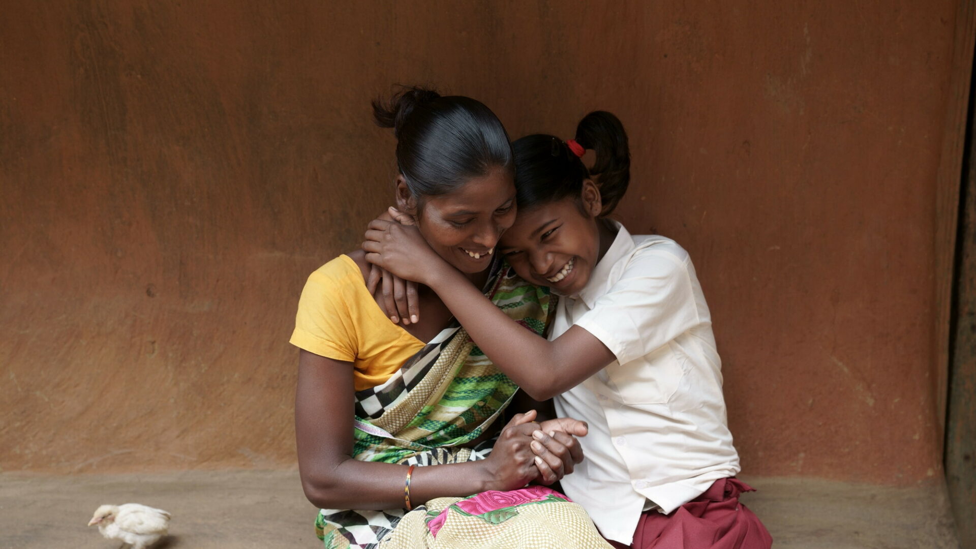 An Indian mother and daughter hug, laughing, while sitting on the step outside the wall of their house