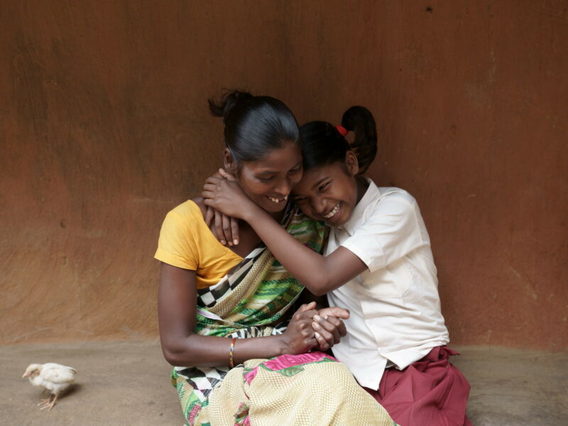 An Indian mother and daughter hug, laughing, while sitting on the step outside the wall of their house