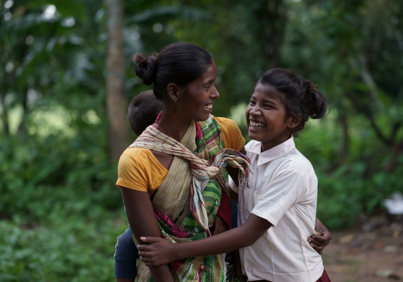 An Indian mother and daughter laugh and hold each other, on a dust road in front of some trees