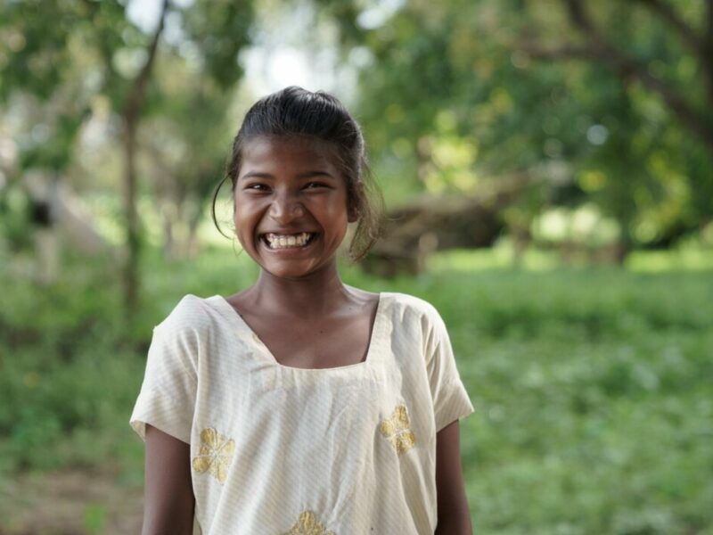 An indian girl in a light coloured blouse stands smiling in the foreground. Behind her is a lush green forest