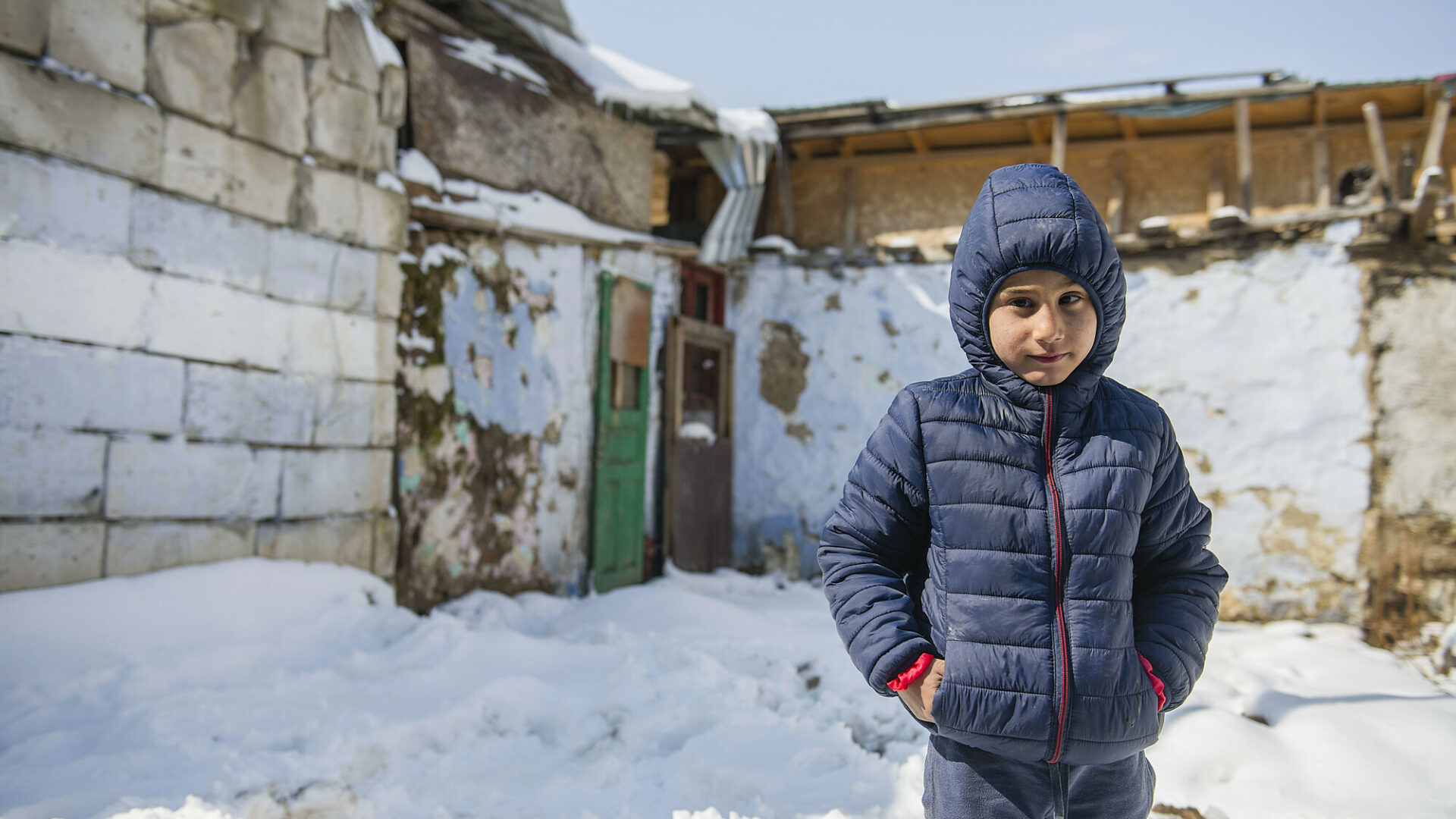 A young romanian boy stands warmly dressed in the snow in front of some farm buildings