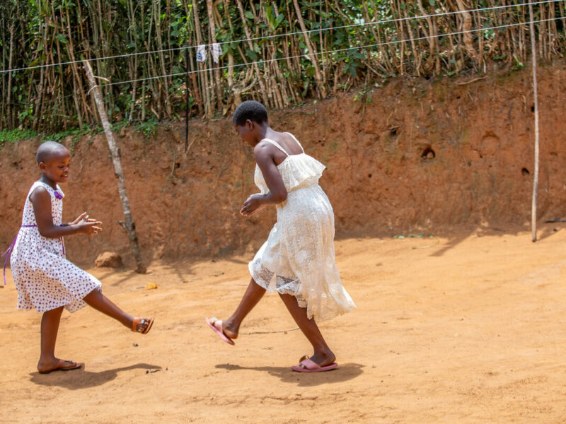 Two Rwandan sisters dance happily together in the red earth backyard of their home
