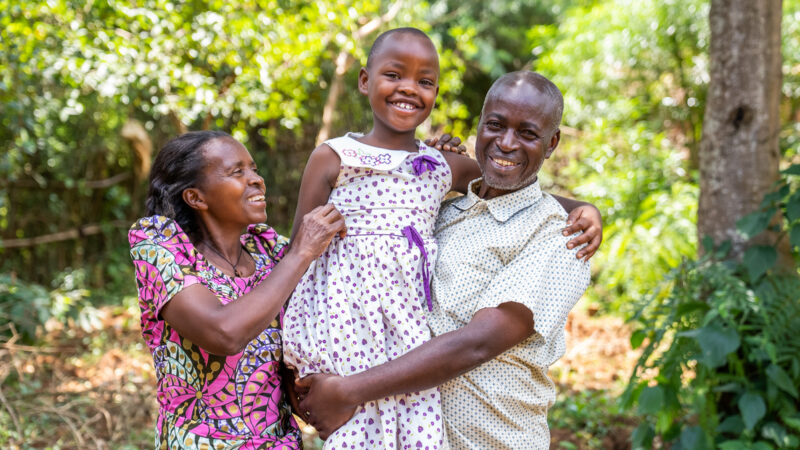 A happy, middle aged Rwandan man holds his 8 year old foster daughter up, smiling at the camera, with his wife on the other side