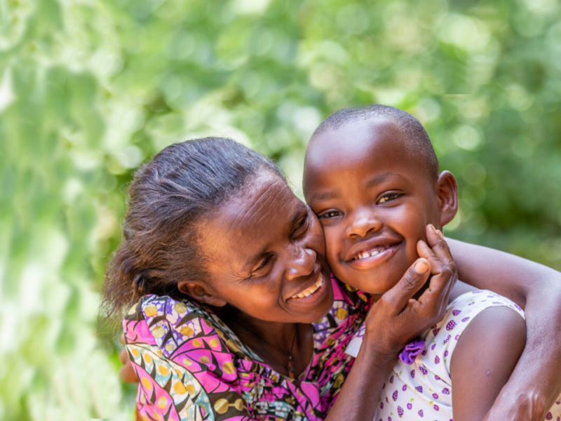 A young Rwandan girl smiles at the camera in front of a leafy green background. Her mother hugs her lovingly.