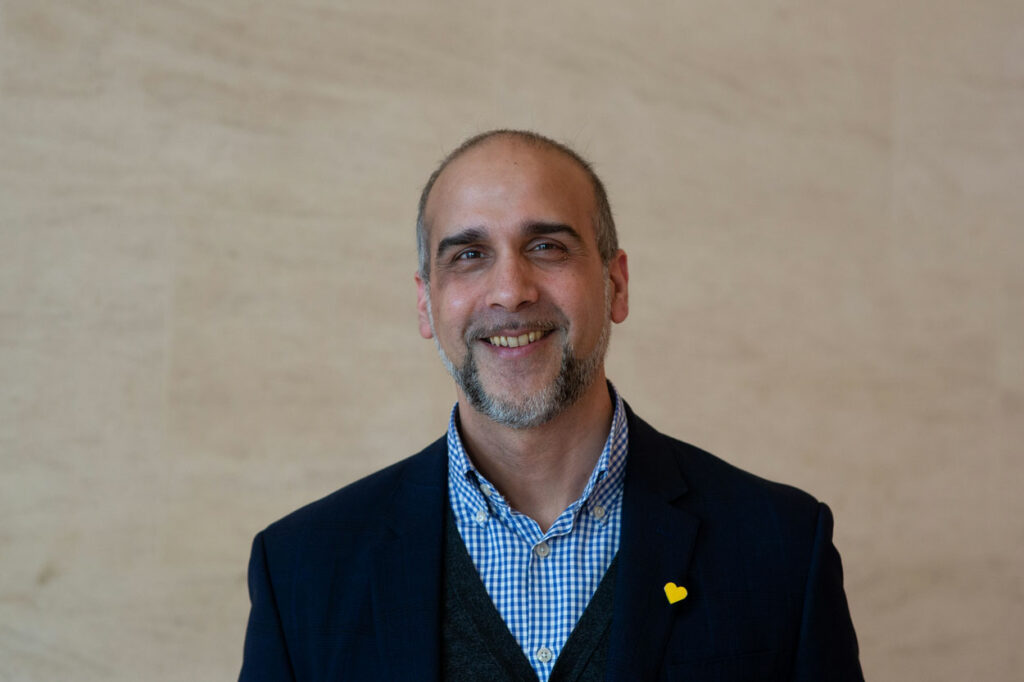 Portrait of Ramesh Parmar, a middle aged man with short grey hair and a beard in a clue check shirt and dark suit jacket - he is smiling at the camera in front of a plain background.