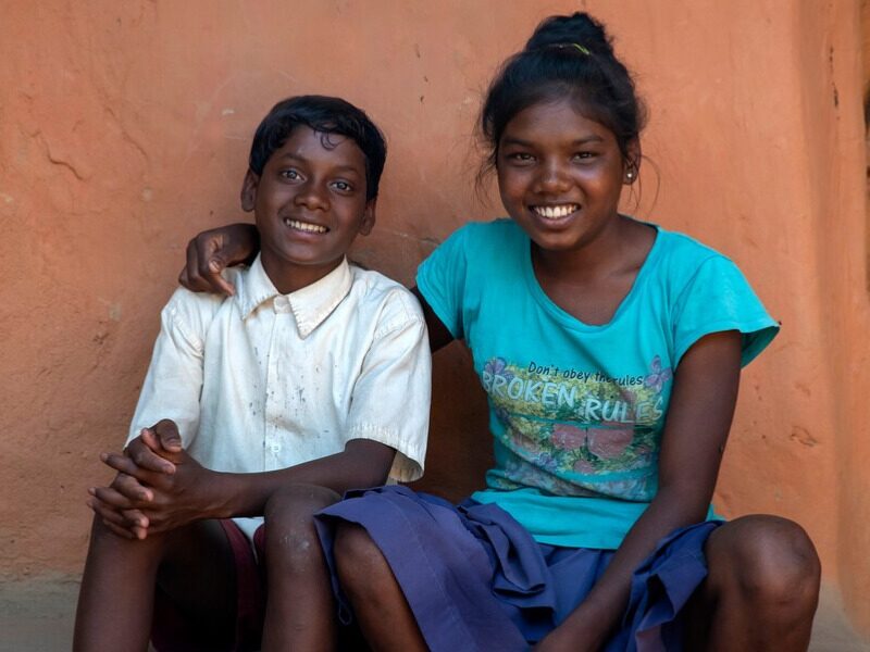 An Indian girl sits with her arm around her younger brother as they both smile at the camera