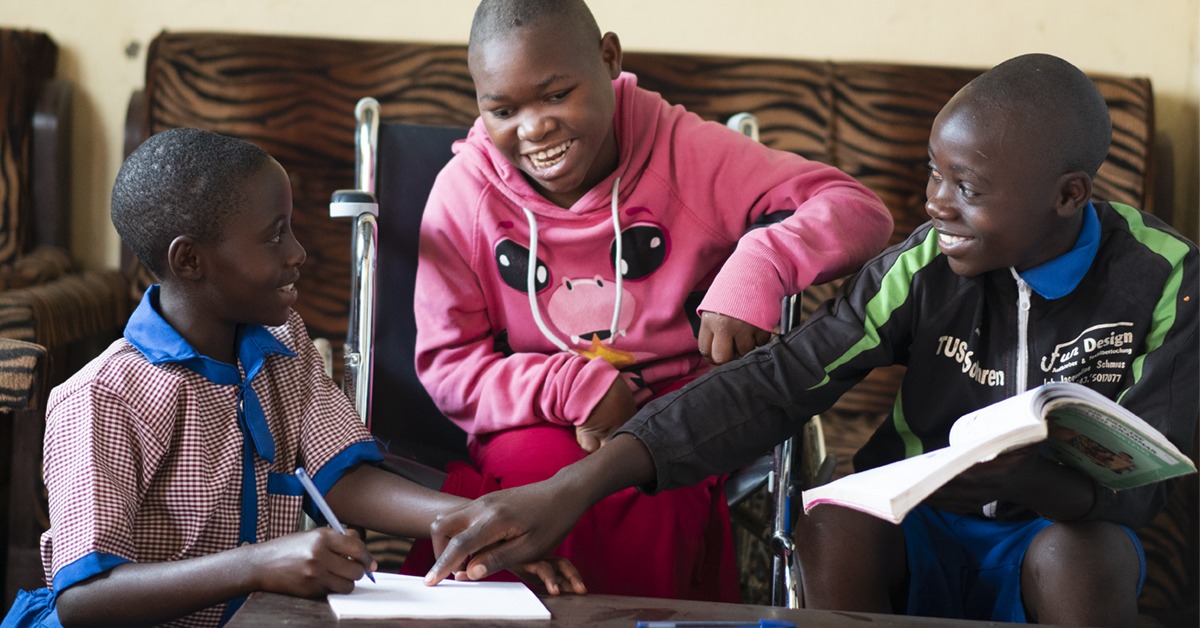 Cary sits in a wheelchair between two other children, one holds a textbook and the other holds a pen to a notepad