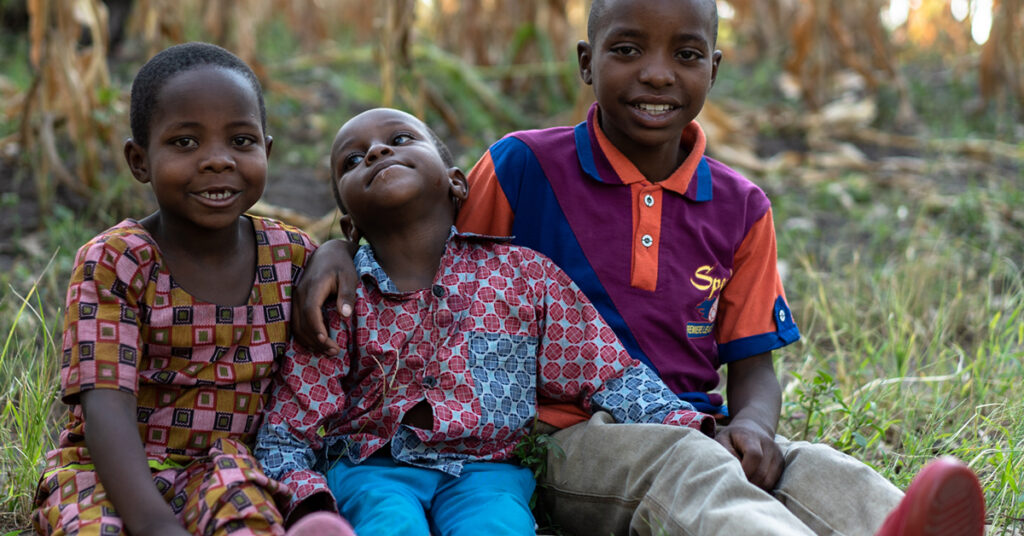 This image is of 3 Rwandan boys (6-9 years old) relaxing together, wearing colourful clothing, sat down on the grass and smiling.