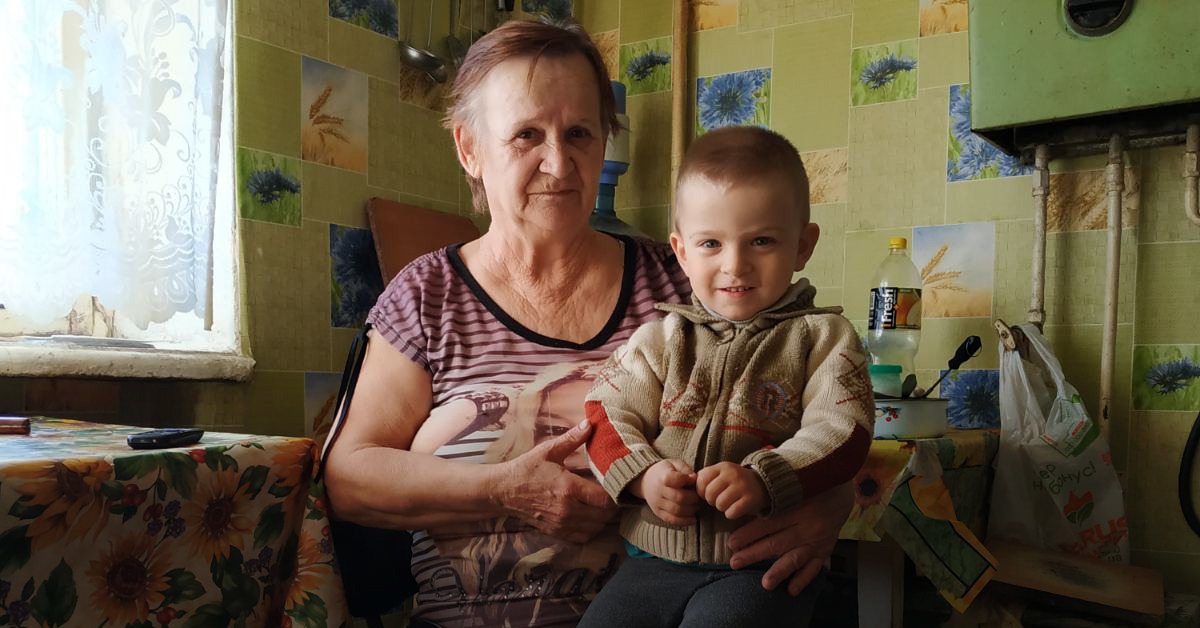 A young Ukrainian boy with short light brown hair, Oleksander, sitting on his grandmother's lap. They look confidently into the camera, in a pale yellow room.