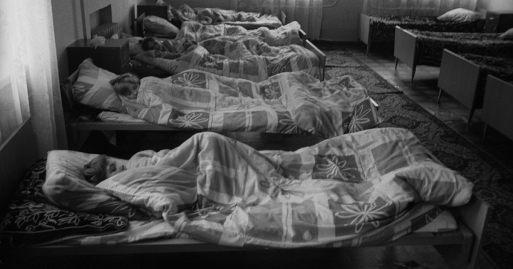 This photo shows a room filled with single beds lined up with children asleep in each one.