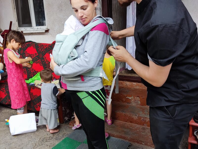 A young dark haired Moldovan woman stands outside her house with her young baby in a carrier. Her partner stands behind her in a green cap, adjusting the straps.