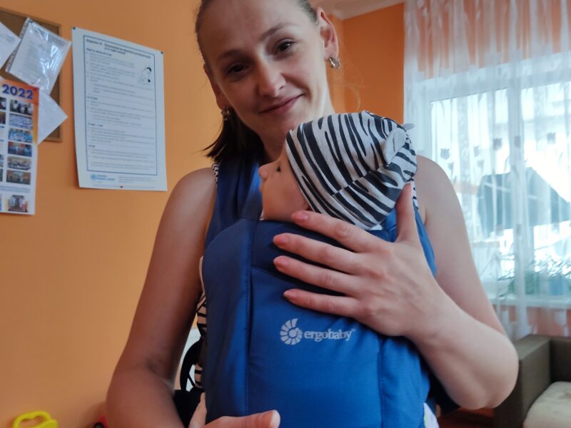 A moldovan woman stands in her orange room, holding a baby on her front in a blue ergobaby carrier. She's smiling at the camera