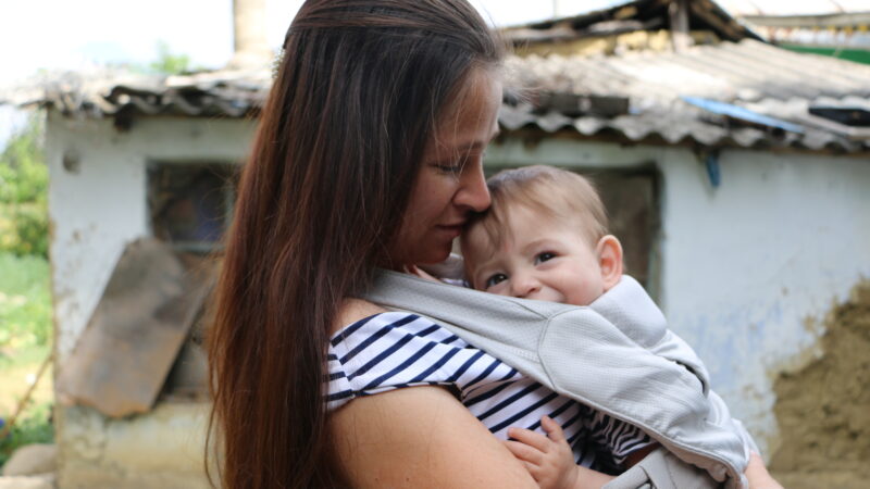 A young Moldovan woman with long dark hair and a stripy top stands with her house in the background, her baby son in a baby carrier on her chest. His head peeps out smiling.