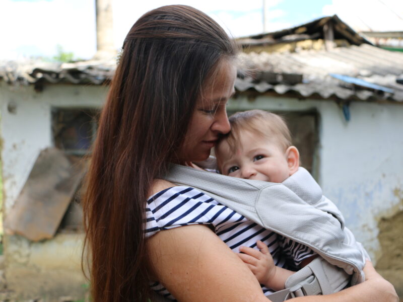 A young Moldovan woman with long dark hair and a stripy top stands with her house in the background, her baby son in a baby carrier on her chest. His head peeps out smiling.