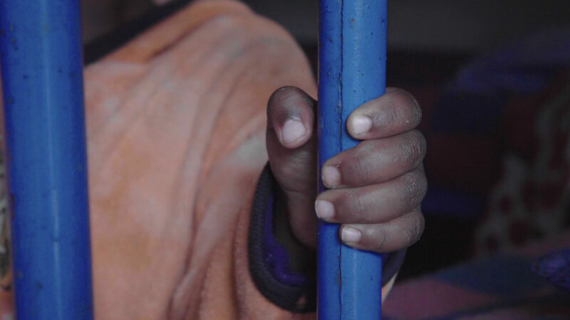 A child's hand holds a painted blue metal bar, locked in a dark orphanage