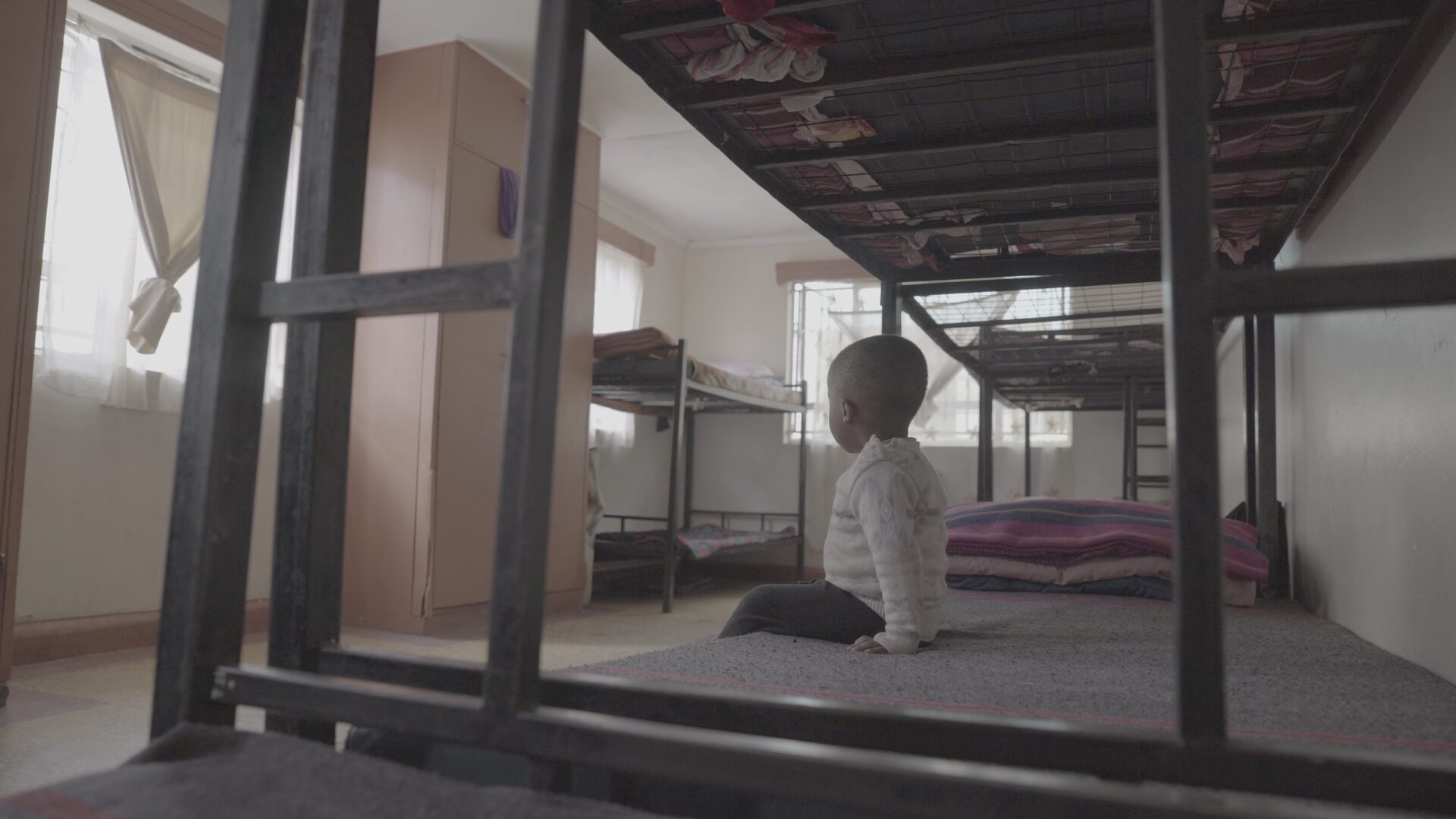 A young Kenyan boy sits alone on his bare metal bunk bed in an empty bleak dormitory