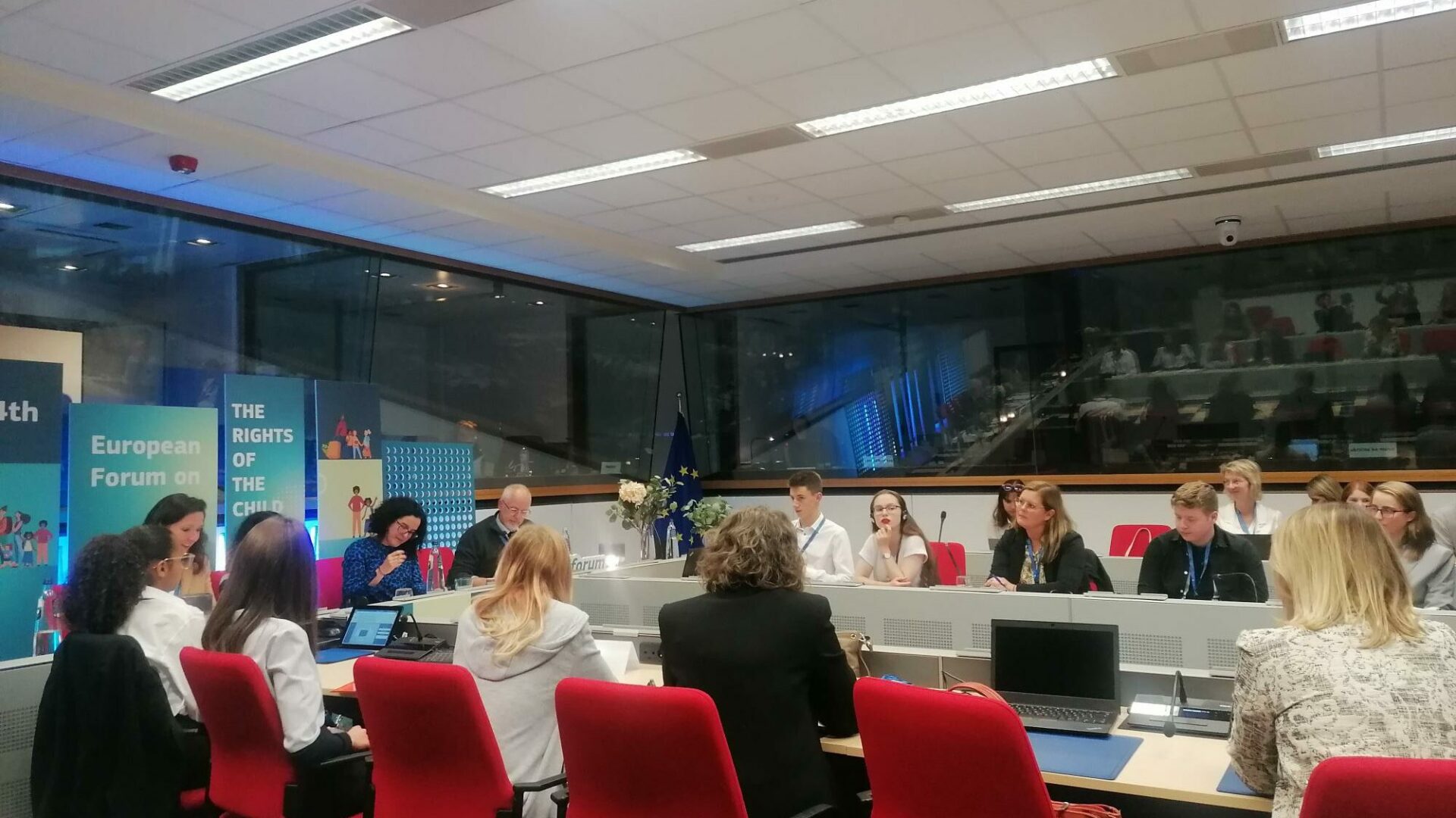 Professionals sat around desks at the EU Child Rights Forum. It's a plain, medium sized office space with large windows on two sides, and 14 professionally dressed men and women sat around a board table with laptops