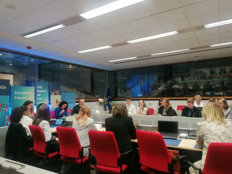 Professionals sat around desks at the EU Child Rights Forum. It's a plain, medium sized office space with large windows on two sides, and 14 professionally dressed men and women sat around a board table with laptops
