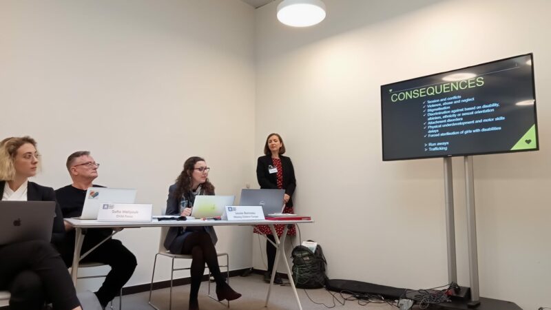 Three professionally dressed men and women sit behind a desk in a plain white office room, while a woman in a suit stands next to a screen on the wall, talking through a presentation.