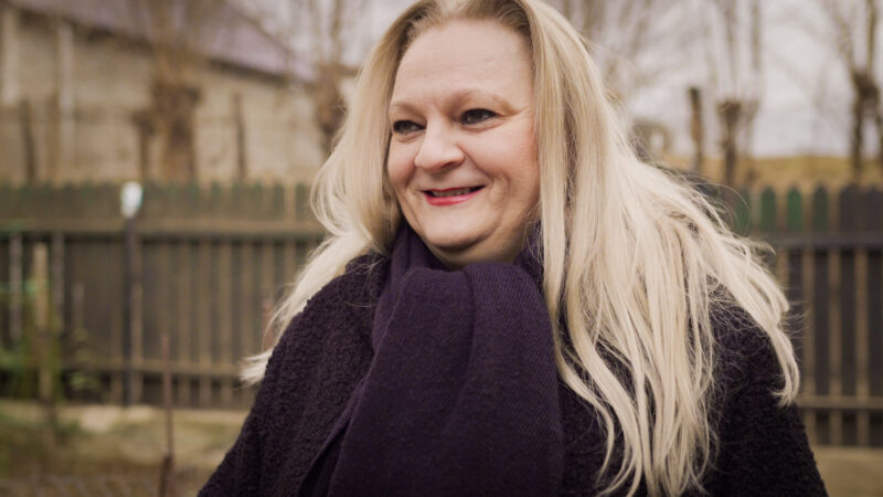 A middle aged white Romanian woman with long blonde hair standing up and smiling in a warm black coat and scarf. She's outside in front of a wooden fence on a dull winter day.