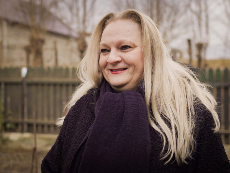 A middle aged white Romanian woman with long blonde hair standing up and smiling in a warm black coat and scarf. She's outside in front of a wooden fence on a dull winter day.