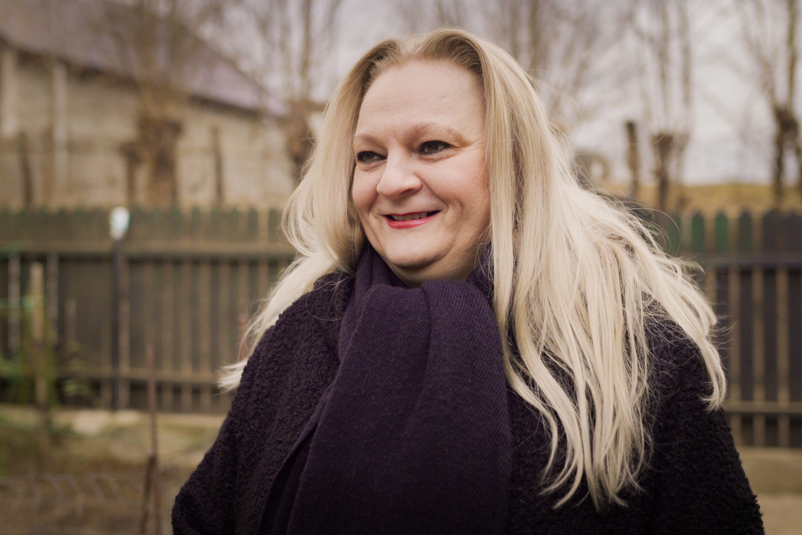 A middle aged white Romanian woman with long blonde hair standing up and smiling in a warm black coat and scarf. She's outside in front of a wooden fence on a dull winter day.