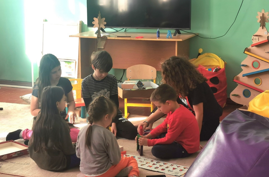 A group of refugee children in a Refugee Accommodation Centre, playing with one of our psychologists in a safe corner set up by our team.