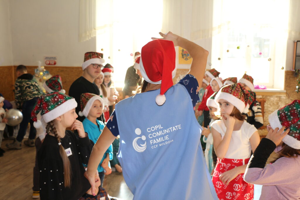 Image is of a person with their back to camera wearing a Santa hat, surrounded by children playing in a circle