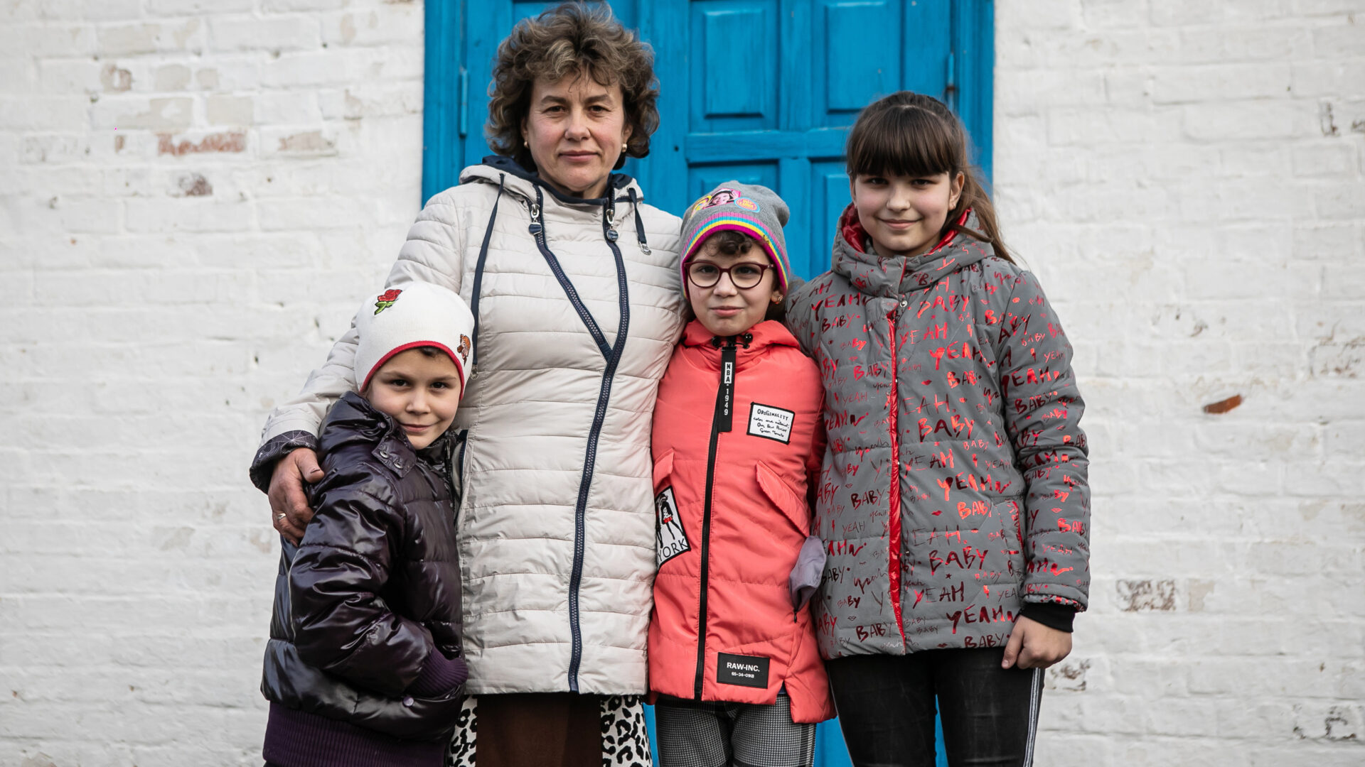 A grandmother, Maria, standing with her arms around her family, a 12 year old girl, and two boys 9 and 7, outside their blue front door. They're all dressed in warm coats, and are smiling