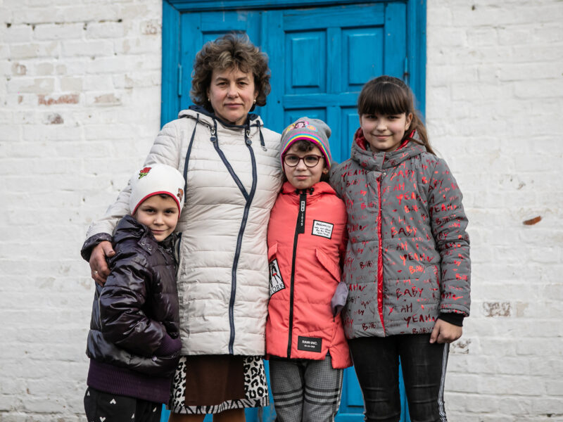 A grandmother, Maria, standing with her arms around her family, a 12 year old girl, and two boys 9 and 7, outside their blue front door. They're all dressed in warm coats, and are smiling