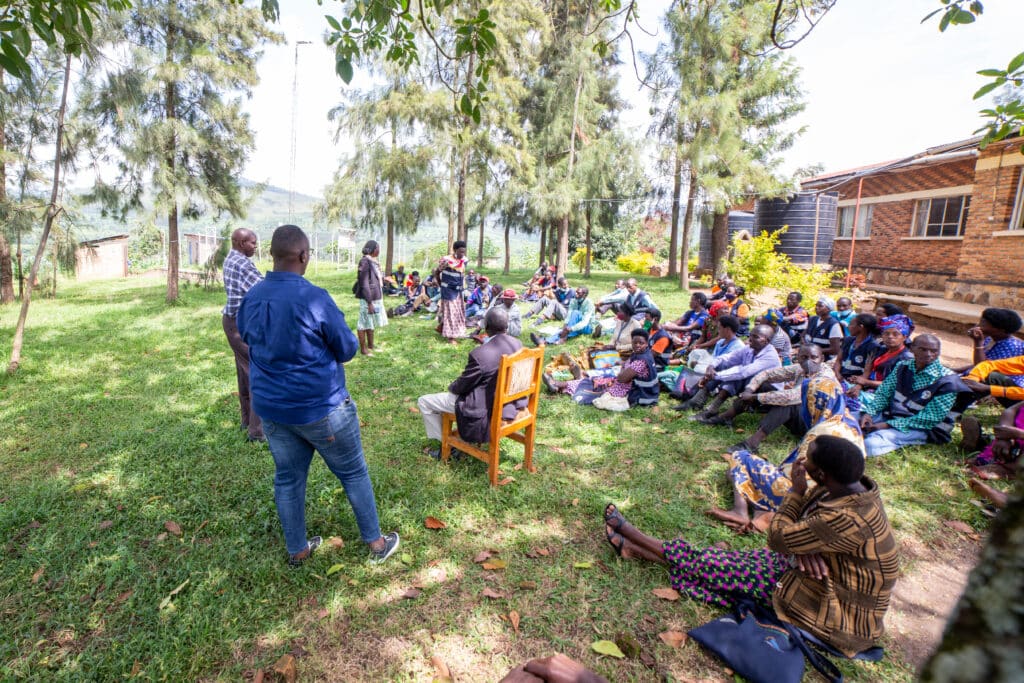 Image is of a meeting, where attendees are sat on the ground outside.