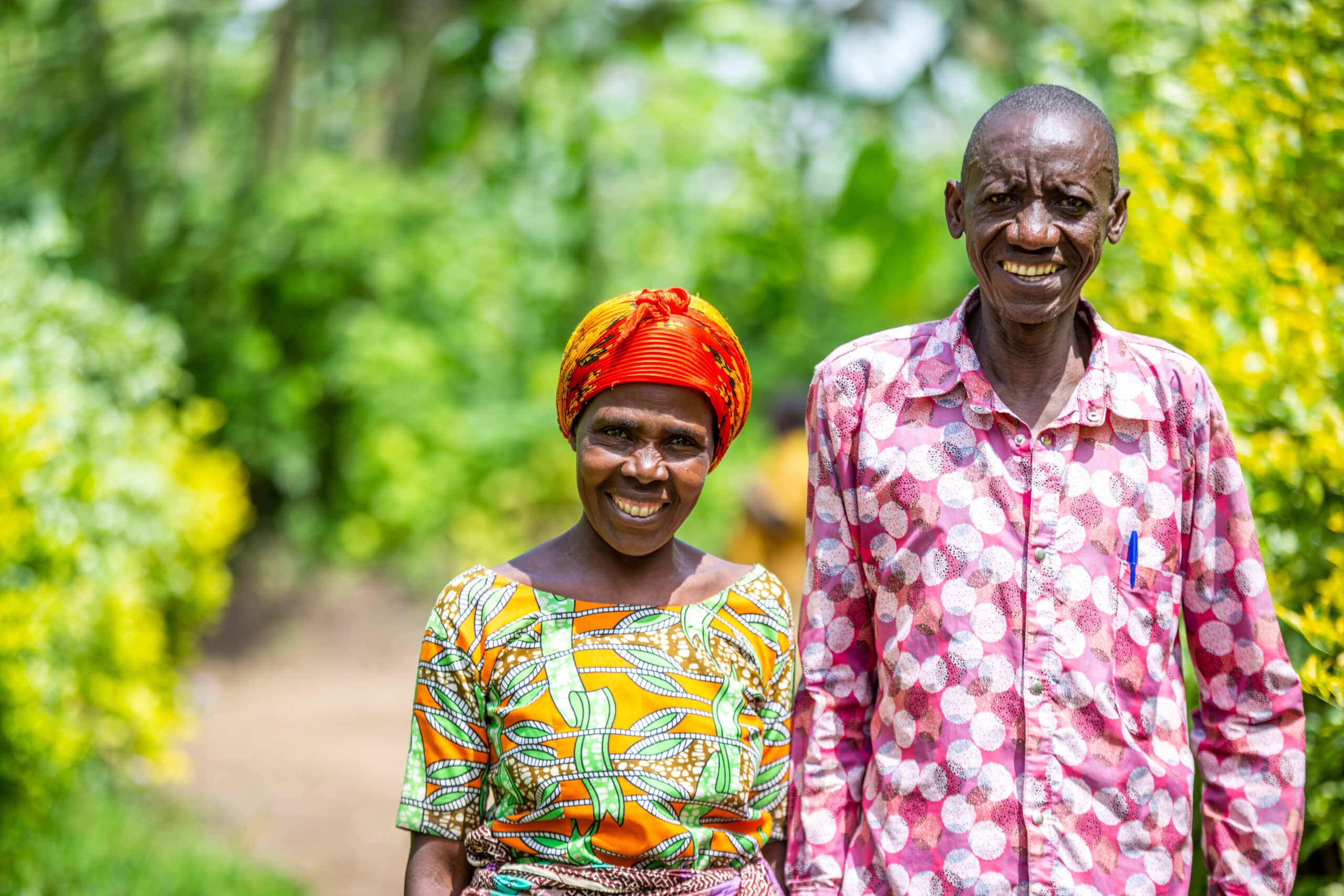 Two older Rwandan foster parents Masaro* and her husband Rugwiro* stand smiling in bright patterned shirts in front of greenery. They look proud and united.